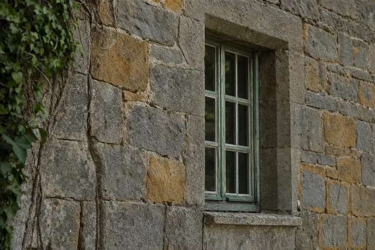 A rough stone wall with a green painted window frame and ivy along one edge